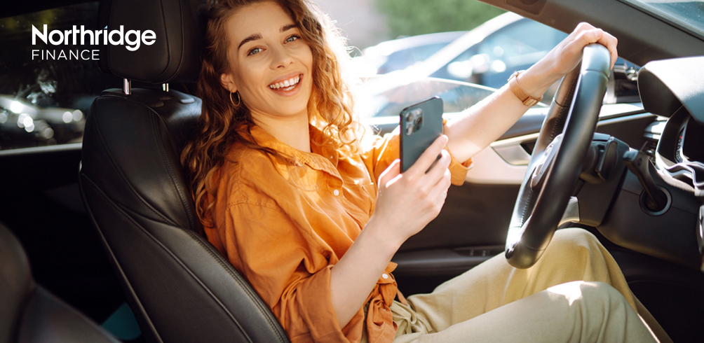 Image of a woman sitting in a car with one hand on the steering wheel and one hand holding her phone in a car showroom